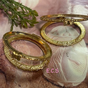 May include: Two gold-toned bangle bracelets with intricate designs. The bracelets are open and have a butterfly-shaped clasp.