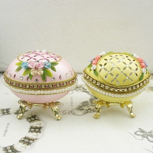 Puede incluir: Dos cajas decorativas en forma de huevo con diseños intrincados. Una caja es rosa con acentos florales y la otra es amarilla con un patrón geométrico. Ambas cajas tienen un borde de perlas y se apoyan en tres patas.