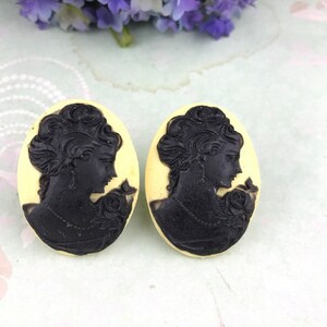 May include: A pair of cream-coloured cameo earrings with black silhouettes of women's profiles. Each earring features a woman with a detailed hairstyle and a floral design.