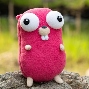Könnte beinhalten: Ein magentafarbenes Plüschtier mit großen, weiß umrandeten Augen und einer kleinen, hellbraunen Schnauze. Das Stofftier hat kleine, abgerundete Arme und Füße. Es sitzt auf einem grauen Felsen mit verschwommenem grünem Hintergrund.