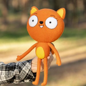 May include: Orange plush cat doll with large white-eyed face, yellow ears, and a yellow belly. The doll has long arms and legs. The doll is being held in front of a blurred background of trees.