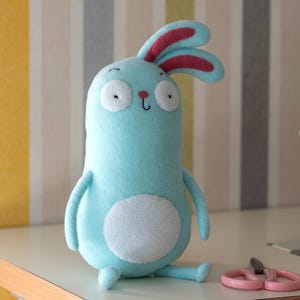May include: A light blue plush bunny with large white eyes and red-lined ears. The handmade stuffed animal has a white belly and is sitting on a white surface. Pink scissors are next to the bunny. The background has vertical stripes.