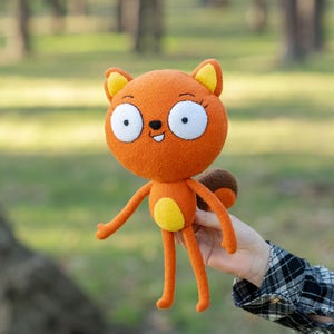 May include: Orange plush squirrel doll with large white eyes, yellow accents, and a brown tail. The doll has long arms and legs and a smiling face. The doll is held in front of a blurred green background.