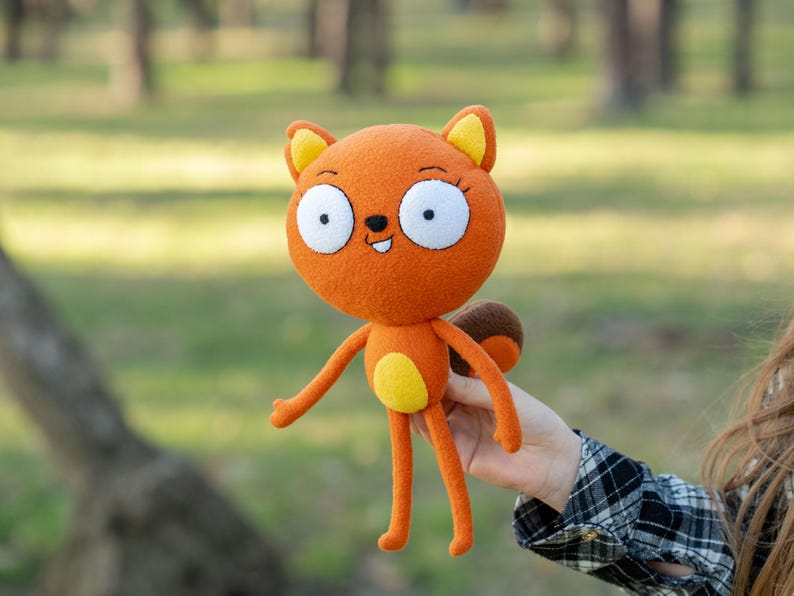 May include: Orange plush squirrel doll with large white eyes, yellow accents, and a brown tail. The doll has long arms and legs and a smiling face. The doll is held in front of a blurred green background.