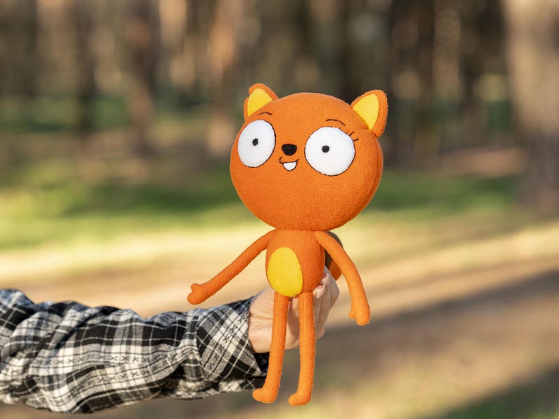 May include: Orange plush cat doll with large white-eyed face, yellow ears, and a yellow belly. The doll has long arms and legs. The doll is being held in front of a blurred background of trees.
