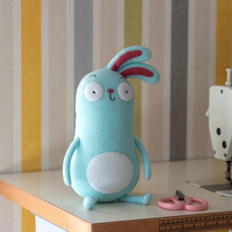 May include: A light blue plush bunny with large white eyes and red-lined ears. The handmade stuffed animal has a white belly and is sitting on a white surface. Pink scissors are next to the bunny. The background has vertical stripes.