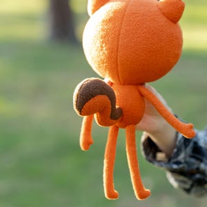 May include: An orange plush fox doll with a rounded head, long legs, and a curved tail. The fox has a brown and orange tail and is held up against a blurred green background.