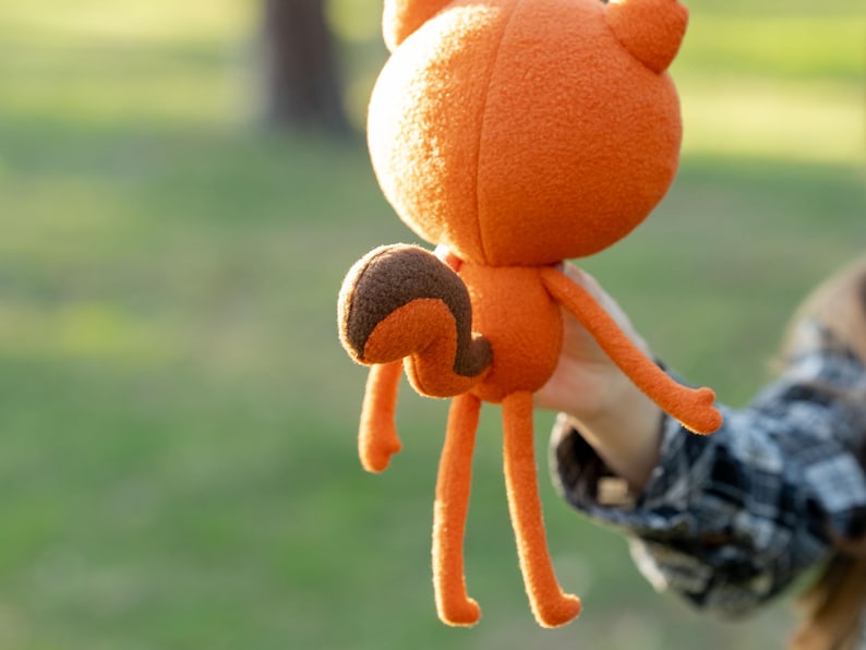 May include: An orange plush fox doll with a rounded head, long legs, and a curved tail. The fox has a brown and orange tail and is held up against a blurred green background.