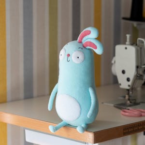 May include: A light blue plush bunny with large white eyes, pink-lined ears, and a white belly patch. The handmade stuffed animal is sitting on a white table with a wooden edge. The background has vertical stripes in yellow, gray, and white.