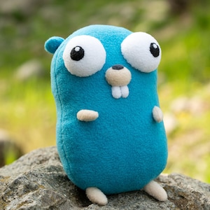 Könnte beinhalten: Ein blaues Stofftier in Form eines gopherartigen Wesens mit großen weißen Augen, einer kleinen Nase und zwei Zähnen. Das Spielzeug hat kleine Arme und Füße und sitzt auf einem Felsen.