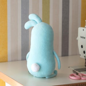 May include: A light blue plush bunny with a white fluffy tail. The handmade stuffed animal has long ears and small arms and legs. The bunny is sitting on a white surface.