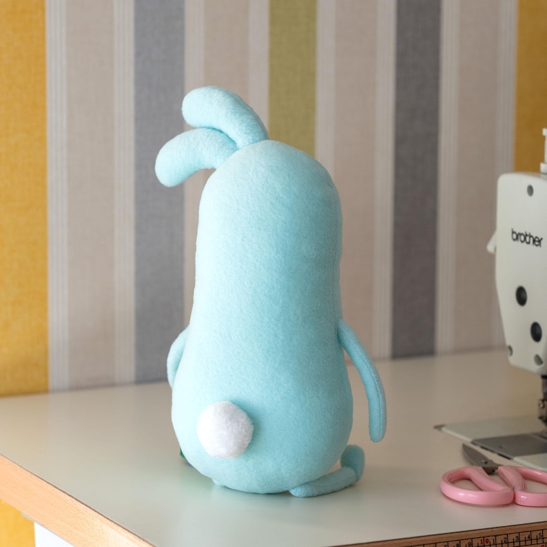 May include: A light blue plush bunny with a white fluffy tail. The handmade stuffed animal has long ears and small arms and legs. The bunny is sitting on a white surface.