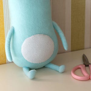 May include: A light blue plush toy with a white circular patch on its back, arms, and legs. Pink scissors are next to the toy. The toy is sitting on a white surface.