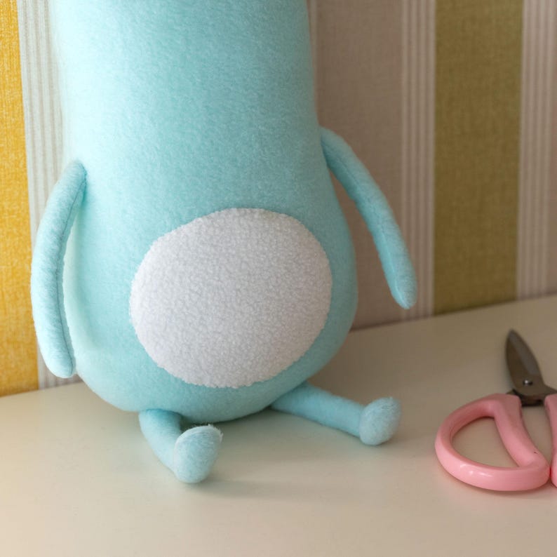 May include: A light blue plush toy with a white circular patch on its back, arms, and legs. Pink scissors are next to the toy. The toy is sitting on a white surface.