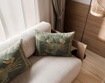 May include: Two rectangular throw cushions in a muted sage green colour, each featuring a detailed, light-coloured graphic print of a scorpion centred within a decorative border. The cushions are placed on a light-coloured, neutral-toned sofa.