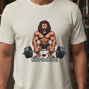 May include: Cream-colored t-shirt featuring a muscular lion lifting a barbell. The lion has a brown mane and is wearing black shorts. The text "Roar in the PR" is below the barbell in a stylized font. The shirt is a crew neck.