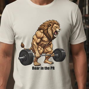 May include: A cream-colored t-shirt featuring a muscular lion lifting a barbell. The lion has a brown mane and is roaring. The text "Roar in the PR" is printed below the lion in black.