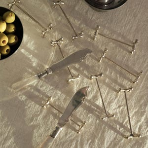 Peut inclure: Vue aérienne d'une table avec des couteaux à beurre argentés et des cure-olives. Les couteaux ont des manches en nacre. Les cure-olives ont un design en forme de croix. Deux plats de service argentés sont en arrière-plan.