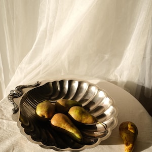 Può includere: Un piatto da portata argentato, smerlato, con un manico ornato, pieno di pere mature. Le pere sono verdi e marroni, e una pera è separata. Il piatto è su una superficie bianca, con una tenda bianca trasparente sullo sfondo.