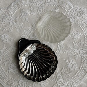 Op de afbeelding: Twee schelpvormige schalen. Eén is van matglas, de andere van glanzend zilvermetaal. Beide hebben een geribbeld ontwerp en liggen op een wit kanten kleedje. De schalen zijn decoratief en kunnen worden gebruikt voor sieraden of kleine items.