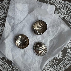 May include: Three silver seashell-shaped dishes on white paper. The dishes have a ribbed texture and a small, round detail at the base. The paper rests on a dark surface with a white lace trim. Each dish is approximately 5 cm in diameter.