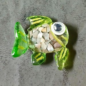 May include: A small fish-shaped ornament crafted from translucent green glass. The body features a mosaic inlay of small, light-colored stone fragments. It has a single white and black googly eye and yellow painted stripes on the fins and tail.