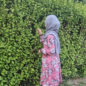 May include: A pink floral tunic with long sleeves and a tiered design. The tunic has a white and grey floral pattern. The person is wearing a grey hijab and brown trousers. The background is a dense green hedge.