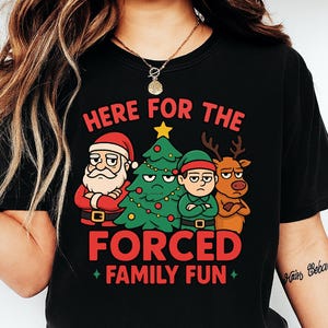 May include: Black t-shirt with a Christmas-themed graphic. The design features Santa Claus, an elf, a Christmas tree, and a reindeer, all with grumpy expressions. The text reads "HERE FOR THE FORCED FAMILY FUN."