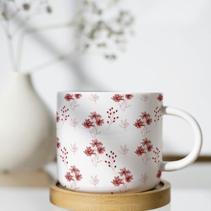 Può includere: Tazza in ceramica bianca con un motivo floreale rosso. La tazza è appoggiata su un sottobicchiere rotondo in legno. Sullo sfondo, un vaso bianco con fiori secchi.