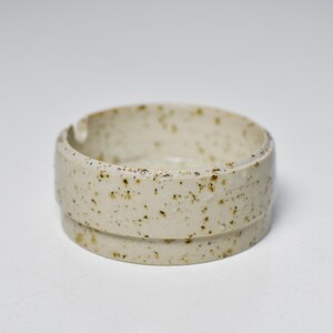 Puede incluir: Cenicero de cerámica beige moteado. Tiene forma circular con una zona rebajada para sujetar cigarrillos. El diseño es sencillo y minimalista, adecuado para uso interior o exterior.