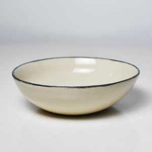 Puede incluir: Un cuenco de cerámica color crema con un borde negro. El cuenco tiene una forma simple y redondeada y un acabado liso y brillante. Este cuenco versátil es adecuado para servir varios platos.