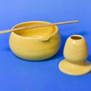 Puede incluir: Un cuenco de cerámica amarilla para matcha con un pequeño pico vertedor, un soporte para batidor de cerámica amarilla a juego y una cuchara de té de bambú larga, fina y curvada que descansa sobre el cuenco. Acabado liso y brillante.