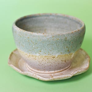 Puede incluir: Un cuenco de cerámica con acabado moteado, que presenta una sección superior de color azul verdoso claro y una sección inferior beige, apoyado sobre un platillo a juego con bordes festoneados. La cerámica tiene una textura con motas marrones.