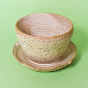 Puede incluir: Un cuenco de cerámica beige moteado con forma redondeada y un interior liso, que descansa sobre un platillo circular a juego con un borde festoneado. La cerámica presenta un esmalte marrón claro con motas marrones más oscuras.