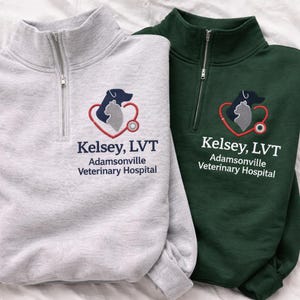 May include: Two quarter-zip sweatshirts, one light gray and one dark green. Each features an embroidered logo of a dog and cat inside a heart with a stethoscope, and the text "Kelsey, LVT, Adamsonville Veterinary Hospital."