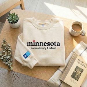 May include: Cream-colored sweatshirt with the word "minnesota" in black script, a red heart, and the text "human decency & hotdish." A small patch with a blue and white design is on the left sleeve. The sweatshirt is on a wooden table.