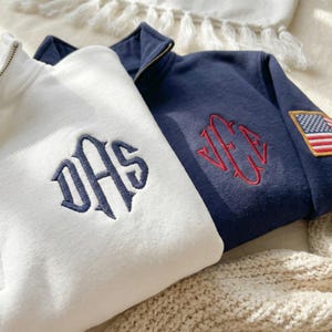 May include: Two fleece pullovers, one white and one navy blue, are displayed. The white pullover has a navy blue embroidered monogram, while the navy blue pullover features a red monogram and an American flag patch. Both have a quarter-zip collar.