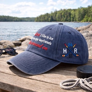 May include: A navy blue baseball cap with the text "ice I like is ice two gay boyfriends around on" embroidered in white and red. The cap also features crossed hockey sticks with the letters "H R" and the numbers "24" and "81".