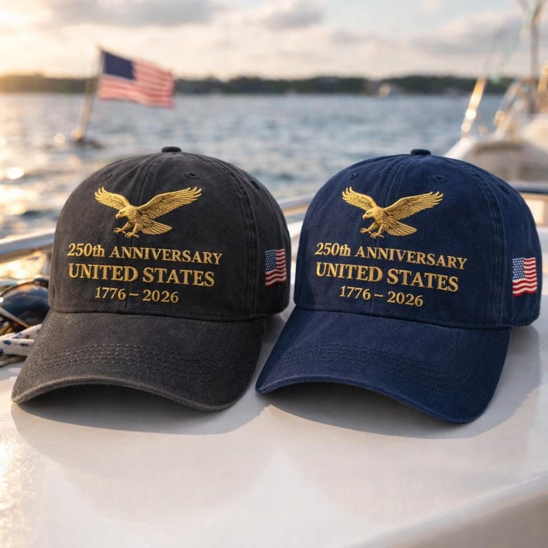 K&ouml;nnte beinhalten: Zwei Baseballkappen, eine schwarze und eine marineblaue, mit goldgesticktem Adler und Text. Der Text lautet "250th ANNIVERSARY UNITED STATES 1776-2026". Jede Kappe hat einen kleinen amerikanischen Flaggenaufn&auml;her. Die Kappen befinden sich auf einem Boot.