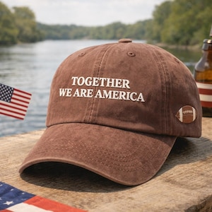 May include: A brown baseball cap with the embroidered text "TOGETHER WE ARE AMERICA" in white. A small football patch is on the side. An American flag is visible in the background.