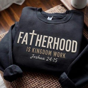 May include: Black crewneck sweatshirt with the embroidered text "FATHERHOOD IS KINGDOM WORK Joshua 24:15" in a light beige color. The sweatshirt is folded on a woven surface, with a white mug visible in the background.