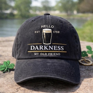 May include: Black baseball cap with the text "HELLO DARKNESS MY OLD FRIEND" and a graphic of a pint of beer. The hat also has the text "EST 1759". The hat is sitting on a stone surface.
