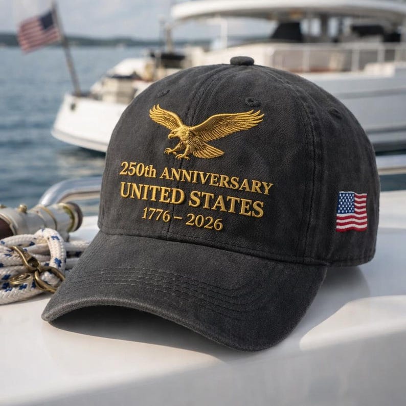 K&ouml;nnte beinhalten: Dunkelgraue Baseballkappe mit goldgesticktem Adler und Text. Der Text lautet "250th Anniversary United States 1776-2026." Auf der Seite befindet sich eine kleine amerikanische Flagge. Die Kappe befindet sich auf einem Boot.