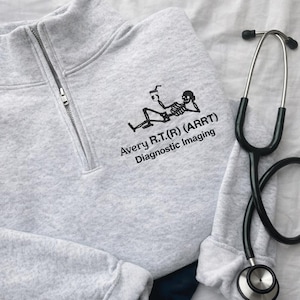 May include: A light gray quarter-zip sweatshirt with the text "Avery R.T.(R) (ARRT) Diagnostic Imaging" and a skeleton graphic. A black stethoscope is next to the sweatshirt.