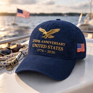 K&ouml;nnte beinhalten: Marineblaue Baseballkappe mit goldgesticktem Text und einem goldenen Adler-Design. Der Text lautet "250th ANNIVERSARY UNITED STATES 1776-2026". Ein kleiner Aufn&auml;her mit der amerikanischen Flagge befindet sich an der Seite. Die Kappe befindet sich auf einem Boot mit einer amerikanischen Flagge im Hintergrund.