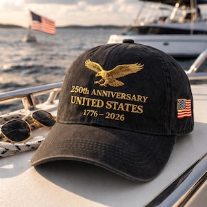 K&ouml;nnte beinhalten: Schwarze Baseballkappe mit goldener Adlerstickerei und dem Text "250th ANNIVERSARY UNITED STATES 1776-2026." Ein amerikanischer Flaggenaufn&auml;her befindet sich an der Seite. Sonnenbrille und ein Boot sind im Hintergrund.