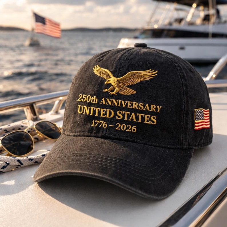 K&ouml;nnte beinhalten: Schwarze Baseballkappe mit goldener Adlerstickerei und dem Text "250th ANNIVERSARY UNITED STATES 1776-2026." Ein amerikanischer Flaggenaufn&auml;her befindet sich an der Seite. Sonnenbrille und ein Boot sind im Hintergrund.