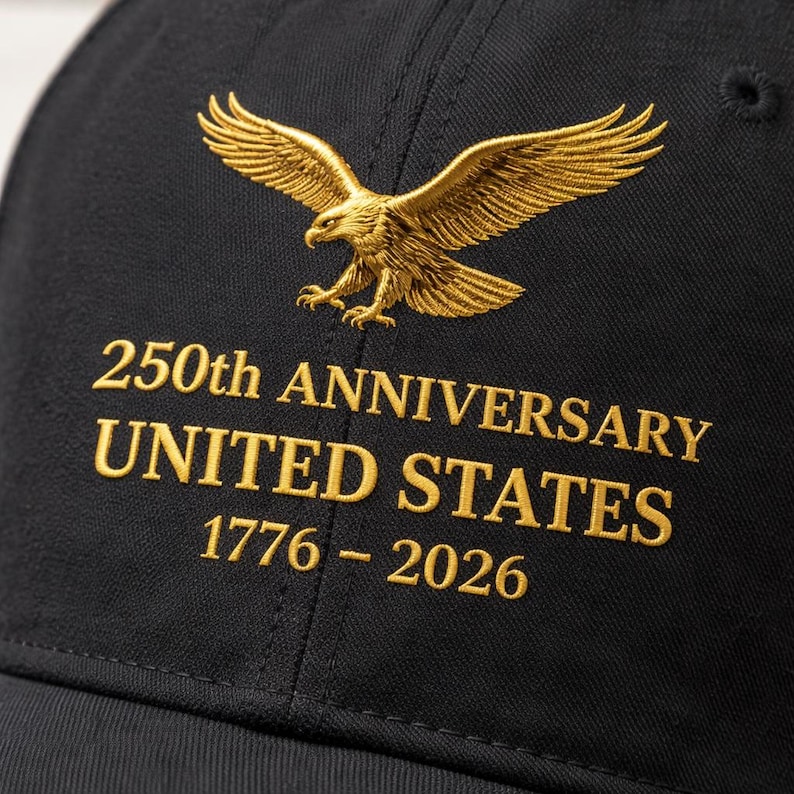 K&ouml;nnte beinhalten: Schwarze Baseballkappe mit goldgesticktem Adler und dem Text: "250th ANNIVERSARY UNITED STATES 1776-2026." Die Kappe hat einen gebogenen Schirm und einen Knopf oben. Der Adler ist mit ausgebreiteten Fl&uuml;geln dargestellt.