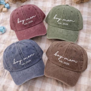 May include: Four baseball caps in shades of burgundy, olive green, denim blue, and brown. Each cap has the text "boy mom est. 2026" in white script across the front. The caps have a vintage, worn look.