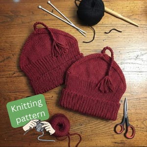 May include: Two knitted burgundy beanies with tassels, displayed on a wooden surface. Knitting needles, black yarn, and scissors are also visible. A green rectangle with the words "Knitting pattern" and a graphic of hands knitting is in the image.